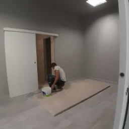 Mężczyzna w szarym pomieszczeniu, klęczący obok wiadra z farbą, przygotowuje płyty kartonowo-gipsowe do montażu, widoczne drzwi przesuwne i szare ściany.