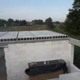 Budowa domu z białych bloczków, widoczne ułożone płyty stropowe z okrągłymi otworami, w tle krajobraz z drogą i zielenią.