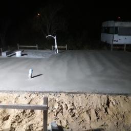 M.S. Vel-BUD Firma Rodzinna - Wylana betonowa płyta fundamentowa pod budynek lub taras, widoczny przewód elektryczny, wiadro i przyczepa kempingowa w tle, nocna sceneria.