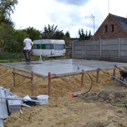 M.S. Vel-BUD Firma Rodzinna - Wylewka betonowa fundamentu pod budynek, widoczny pracownik w kaloszach, przyczepa kempingowa w tle i narzędzia budowlane.