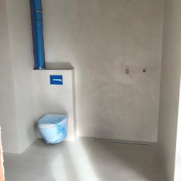 Mkcement - Łazienka w trakcie wykończenia z szarymi ścianami i podłogą, toaleta zabezpieczona folią, widoczne rury i otwory pod armaturę.