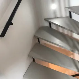 Schody z szarego mikrocementu z czarną metalową balustradą zamontowane przy białej ścianie z oświetleniem punktowym.