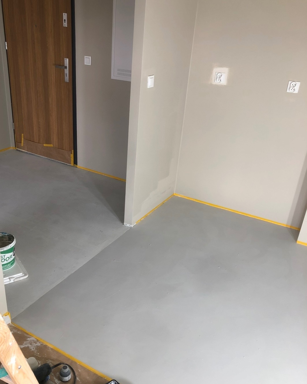 Świeżo wylana, szara posadzka w mieszkaniu, zabezpieczona żółtą taśmą malarską przy ścianach, widoczny kubeł z resztkami produktu i narzędzia pracy.