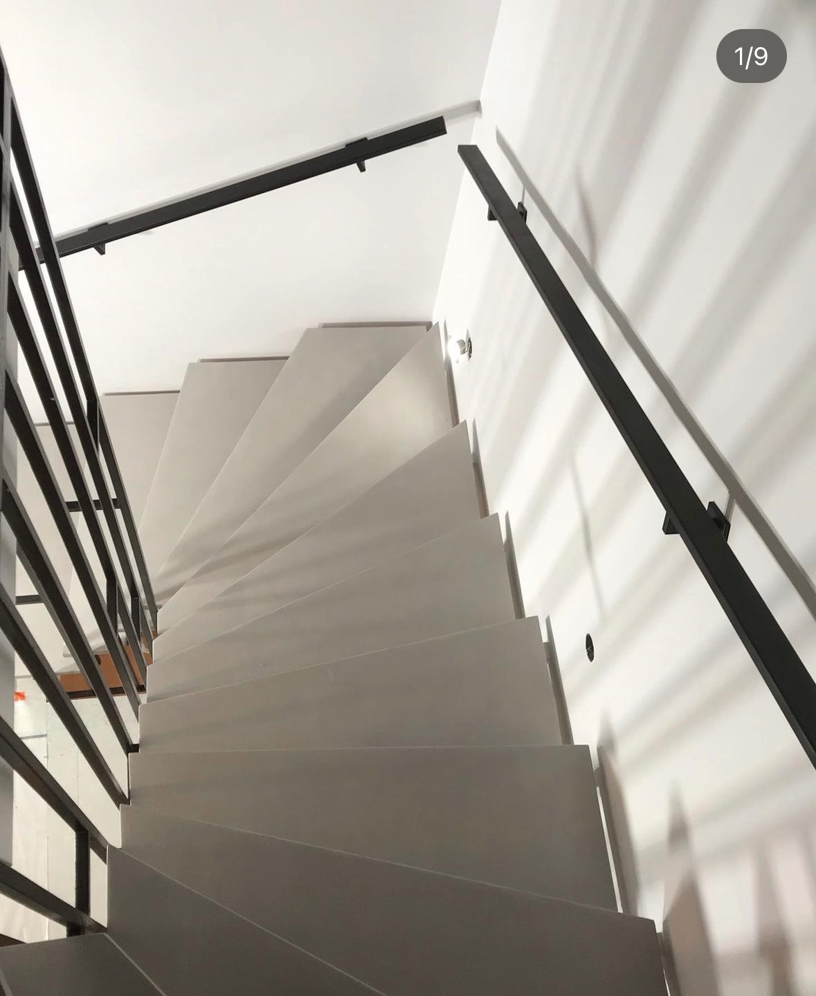 Widok z góry na białe, minimalistyczne schody z czarną metalową balustradą i poręczą, oświetlone naturalnym światłem, z widocznym numerem zdjęcia 1/9 w prawym górnym rogu.