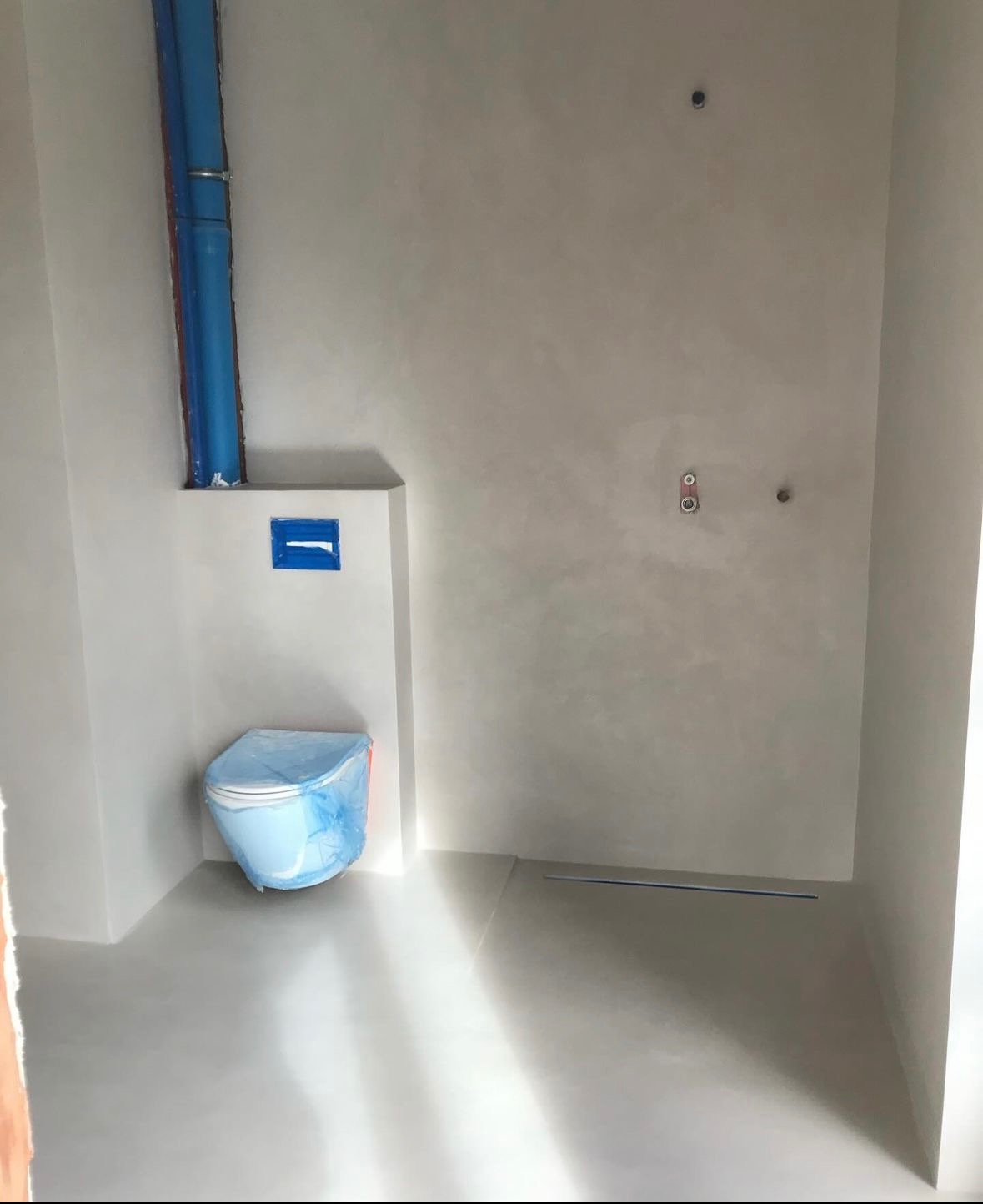 Łazienka w trakcie wykończenia z szarymi ścianami i podłogą, toaleta zabezpieczona folią, widoczne rury i otwory pod armaturę.