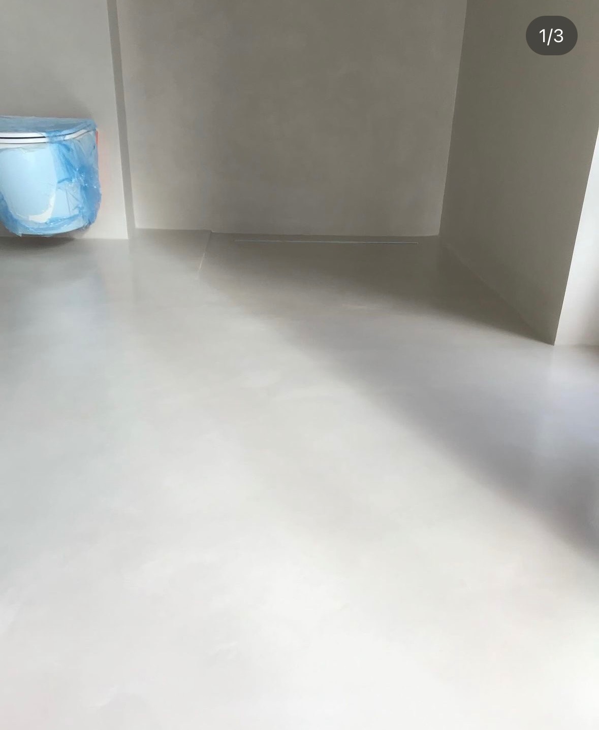 Jasnoszara, błyszcząca posadzka z mikrocementu w minimalistycznej łazience, toaleta zabezpieczona niebieską folią ochronną.