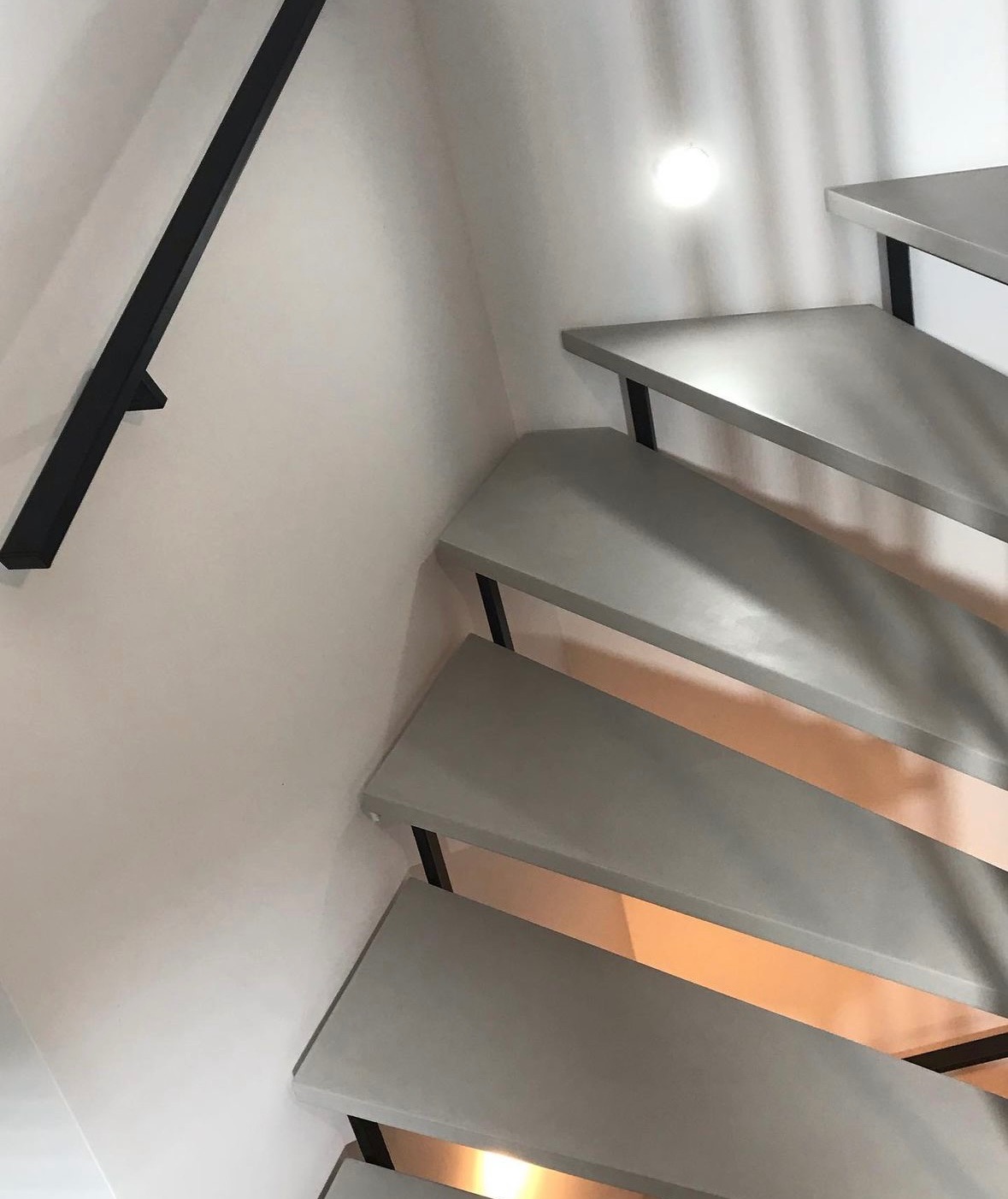 Schody z szarego mikrocementu z czarną metalową balustradą zamontowane przy białej ścianie z oświetleniem punktowym.