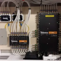 Rozdzielnica telekomunikacyjna marki Televes z licznymi kablami i złączami, zamontowana w białej szafce, widoczne etykiety na kablach.