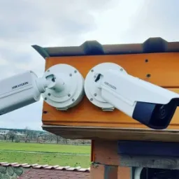 Dwie kamery monitoringu Hikvision zamontowane na drewnianej elewacji budynku, widok na zielony krajobraz w tle.