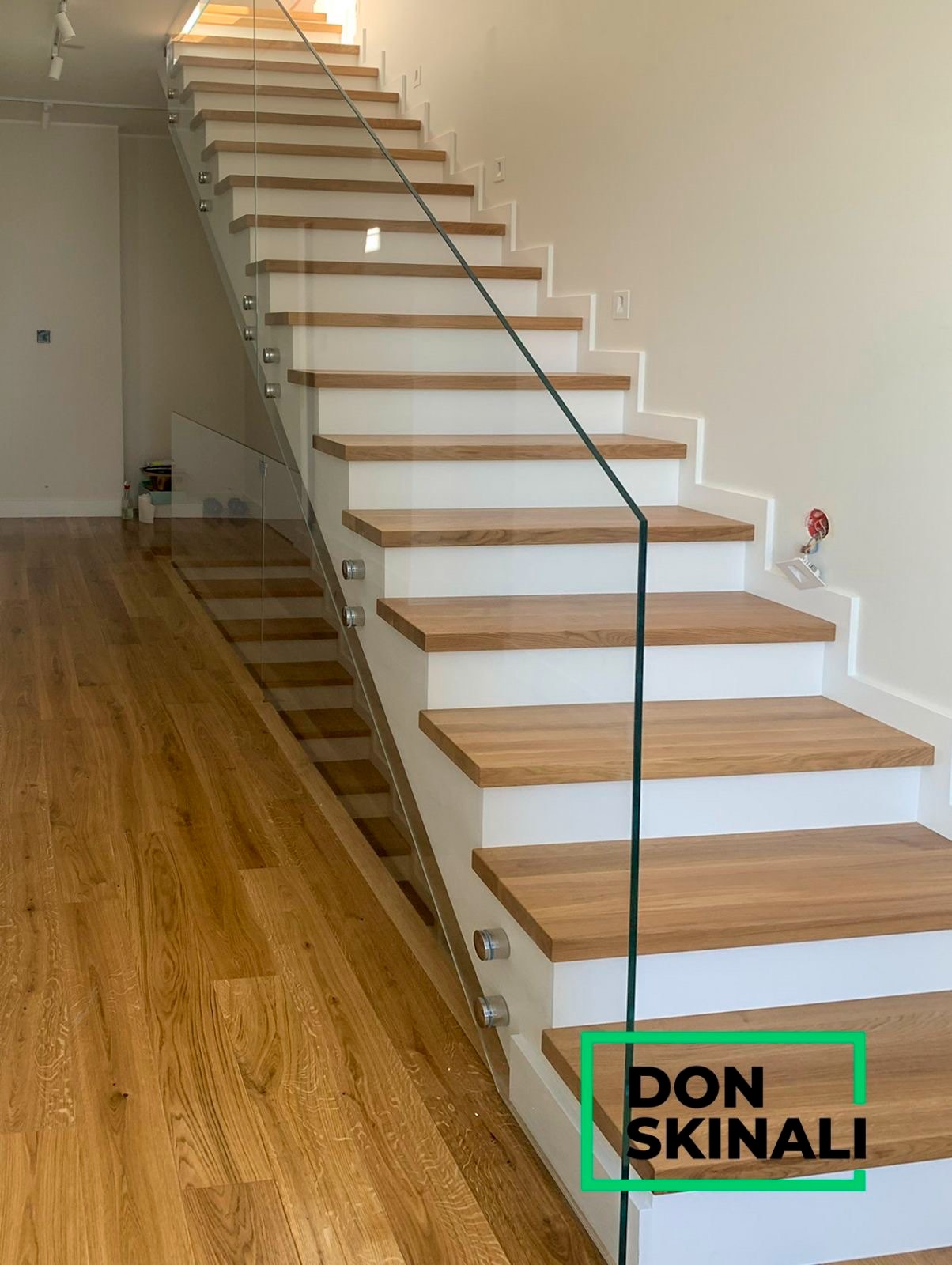 Nowoczesne schody z drewnianymi stopniami i szklaną balustradą mocowaną na metalowych uchwytach, widoczne z boku, oświetlone naturalnym światłem.