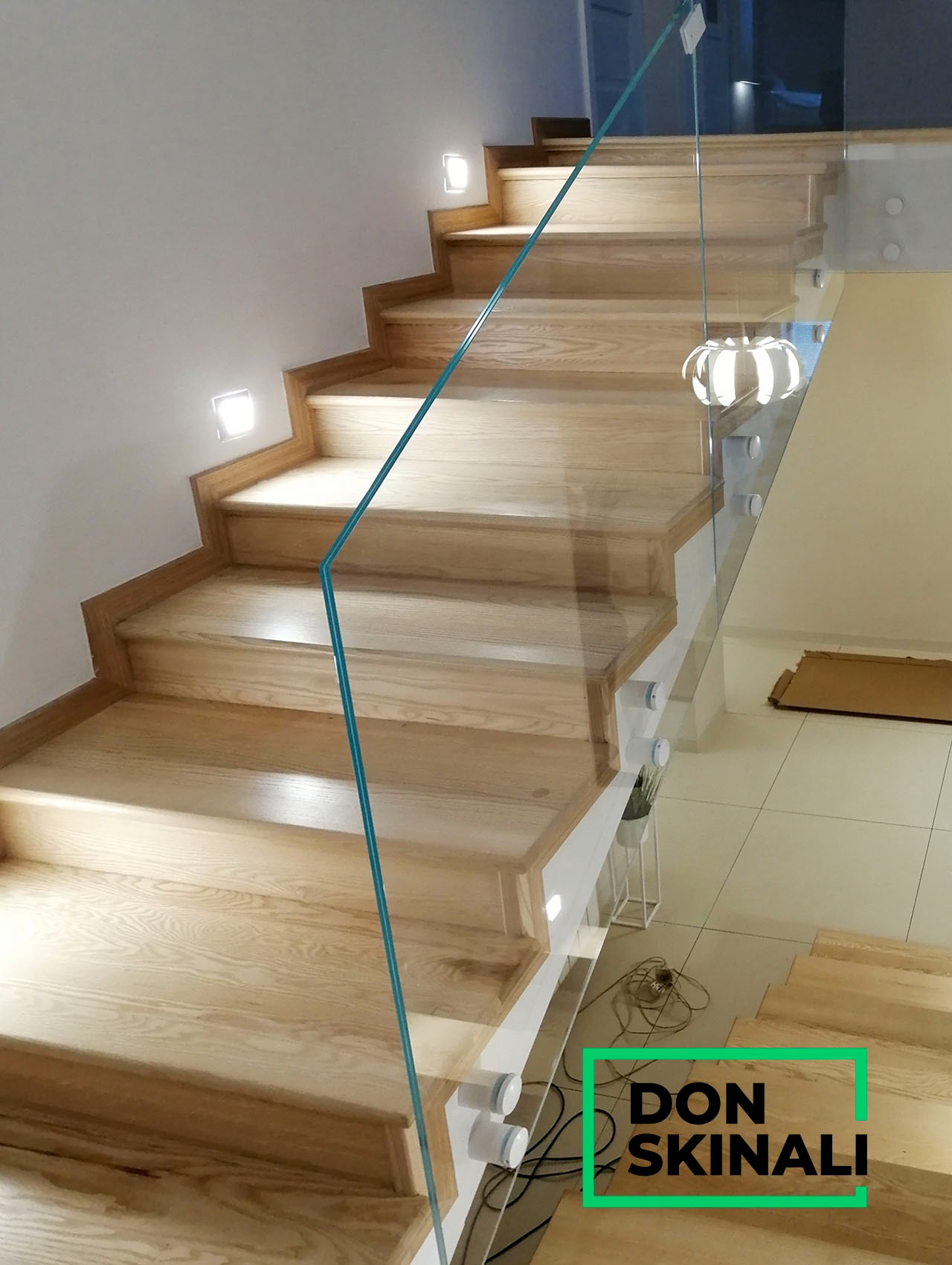 Schody drewniane z balustradą szklaną mocowaną na punktowych uchwytach, oświetlenie schodowe wbudowane w ścianę, widoczny napis 'DON SKINALI'.
