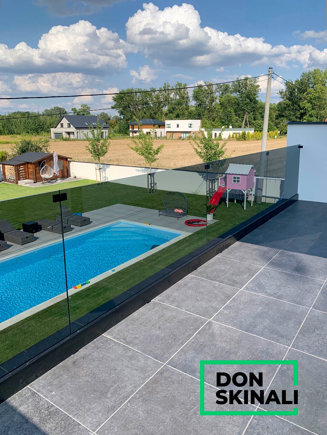 Nowoczesny ogród z basenem, widok z tarasu zabezpieczonego szklaną balustradą, w tle domek dla dzieci i budynki mieszkalne.