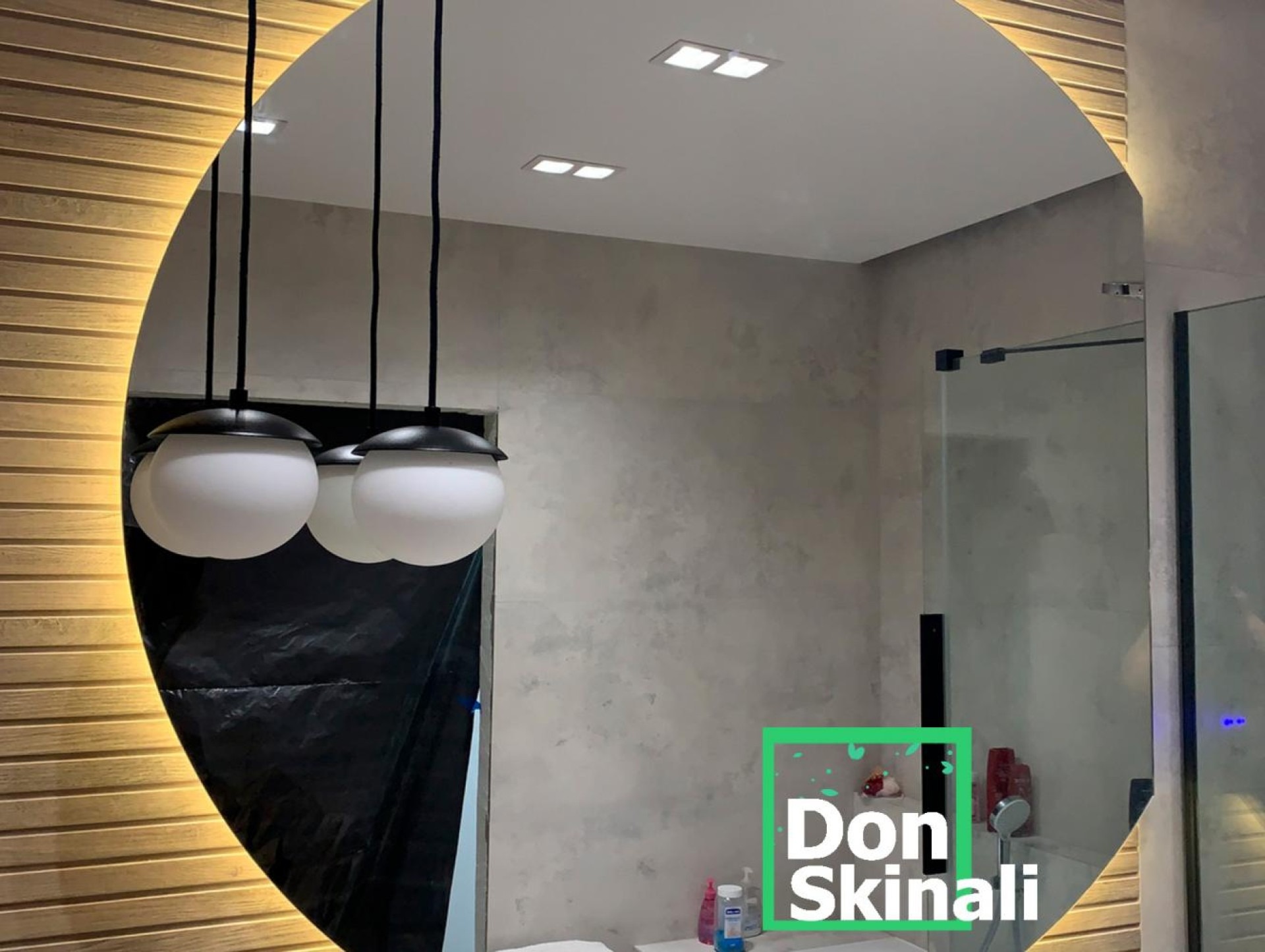 Owalne lustro z podświetleniem LED, odbijające nowoczesną łazienkę z kabiną prysznicową typu walk-in, betonową ścianą i wiszącymi lampami kulami. Fragment czarnej folii ochronnej widoczny w dolnej...