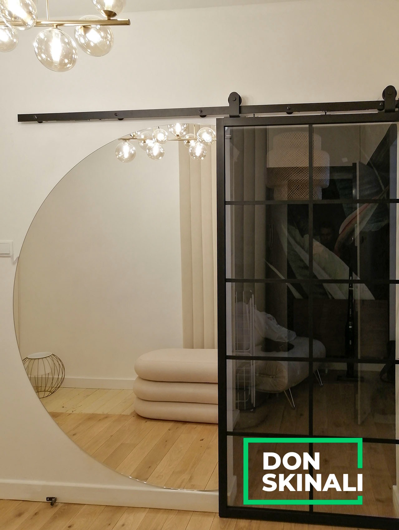 Szklane drzwi przesuwne w czarnej ramie z widocznym logo firmy Don Skinali, zamontowane na szynie, odbijające się w okrągłym lustrze, z widocznym oświetleniem sufitowym i beżową pufą.
