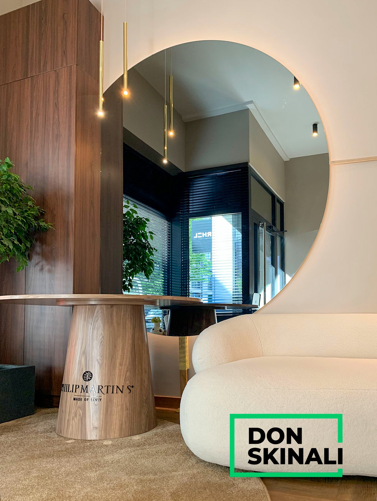 Nowoczesne wnętrze z okrągłym lustrem z podświetleniem LED, drewnianym stołem z logo i jasną sofą w tle okna z żaluzjami.