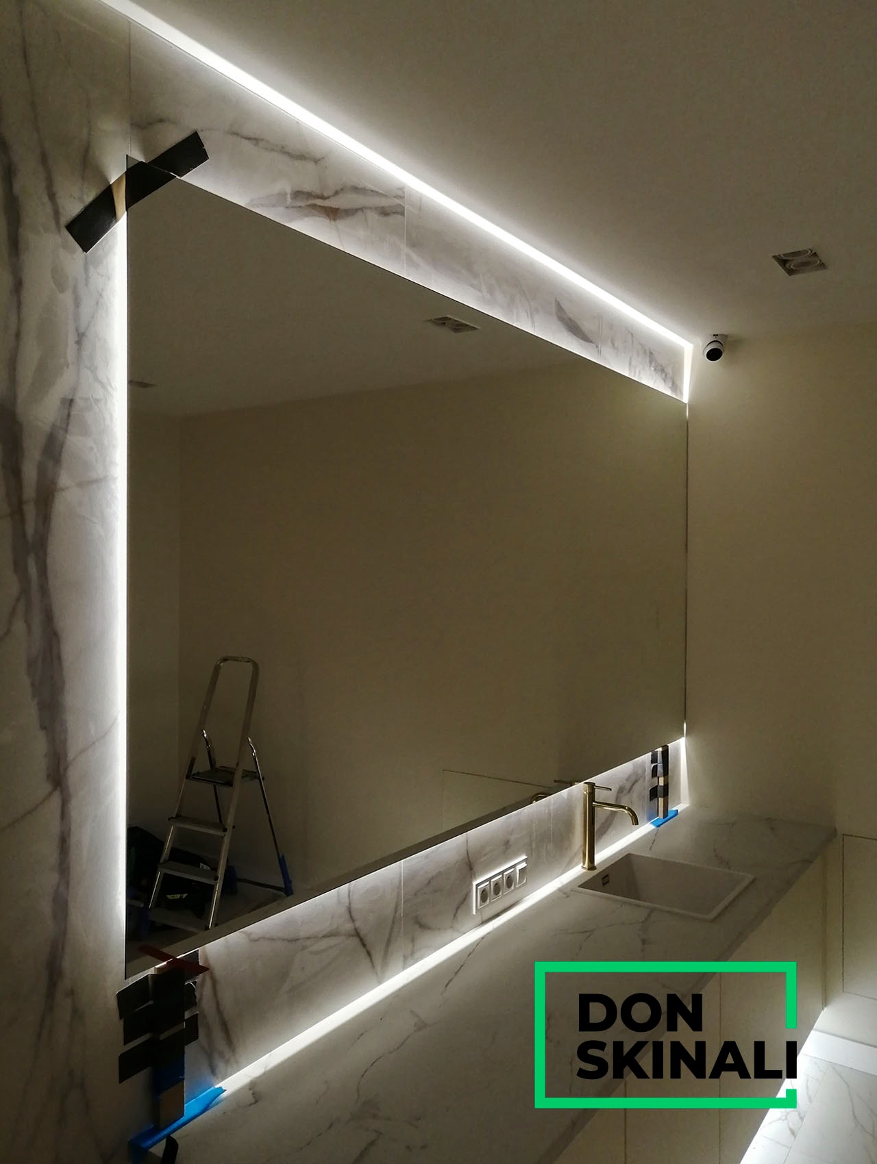 Duże lustro łazienkowe z podświetleniem LED, obudowane marmurowymi płytami, widoczna drabina i elementy montażowe, złota bateria umywalkowa.