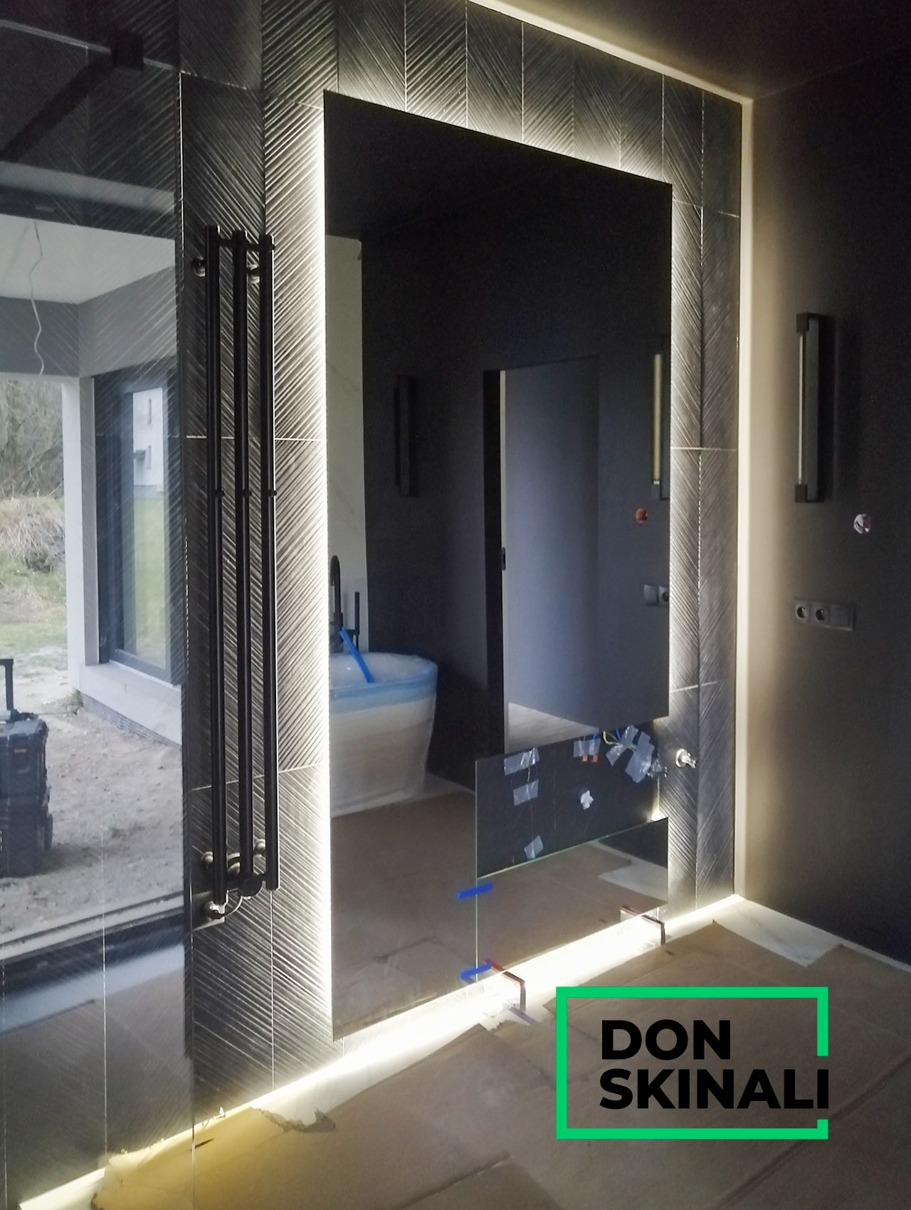 Szklana ściana z podświetleniem LED i czarnym grzejnikiem drabinkowym, widok na łazienkę w trakcie remontu, z zabezpieczoną folią wanną i wystającymi kablami.