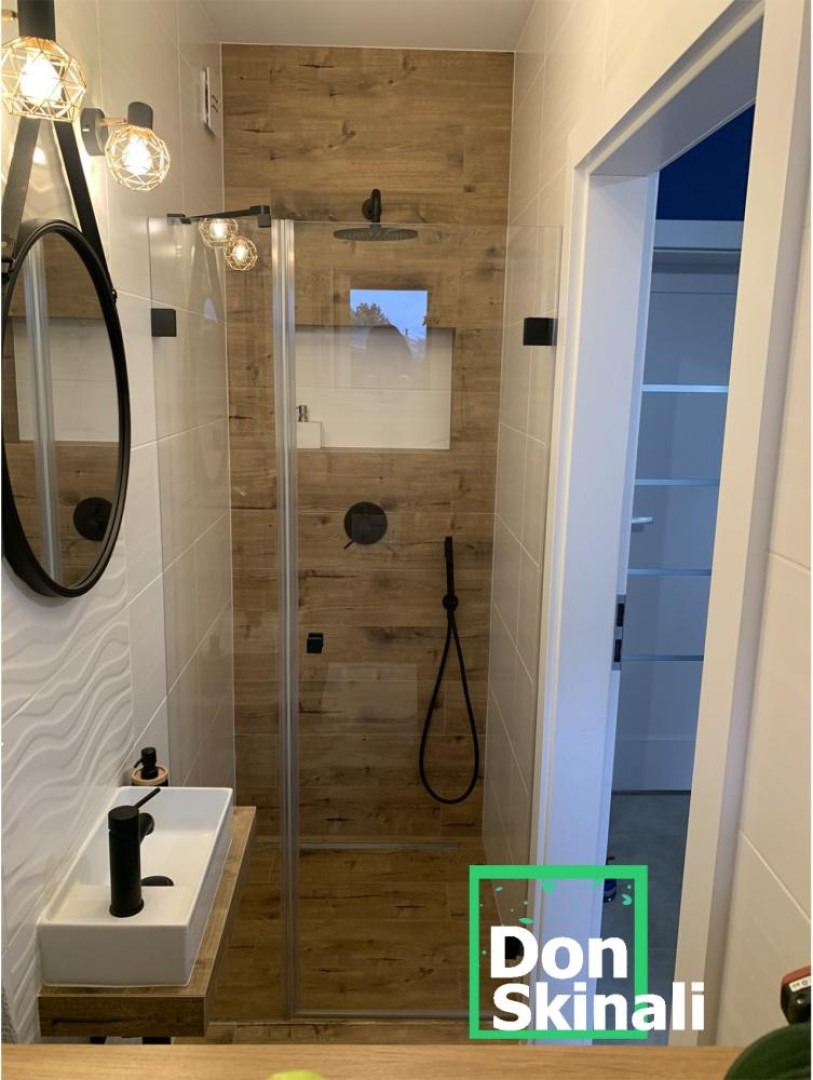 Nowoczesna łazienka z kabiną prysznicową ze szklanymi ściankami, wykończona płytkami imitującymi drewno, z czarną armaturą i owalnym lustrem na ścianie z dekoracyjną fakturą.