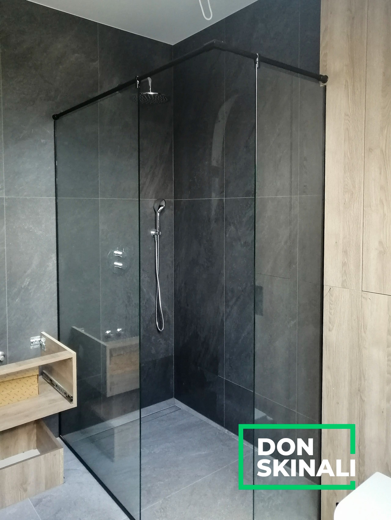 Nowoczesna kabina prysznicowa walk-in z czarnymi profilami, szklana ściana, szara glazura w łazience, widoczny fragment szafki pod umywalkę z otwartymi szufladami.