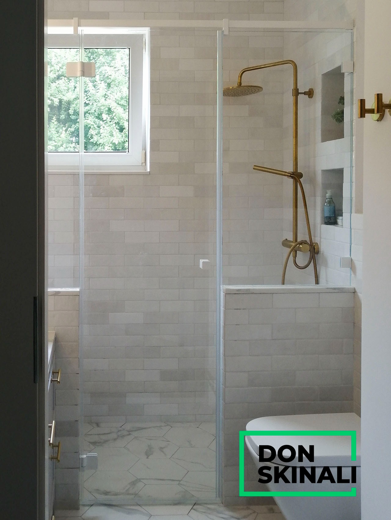 Szklana kabina prysznicowa z złotą armaturą i deszczownicą w łazience w Warszawie, widok na okno i heksagonalne płytki podłogowe.