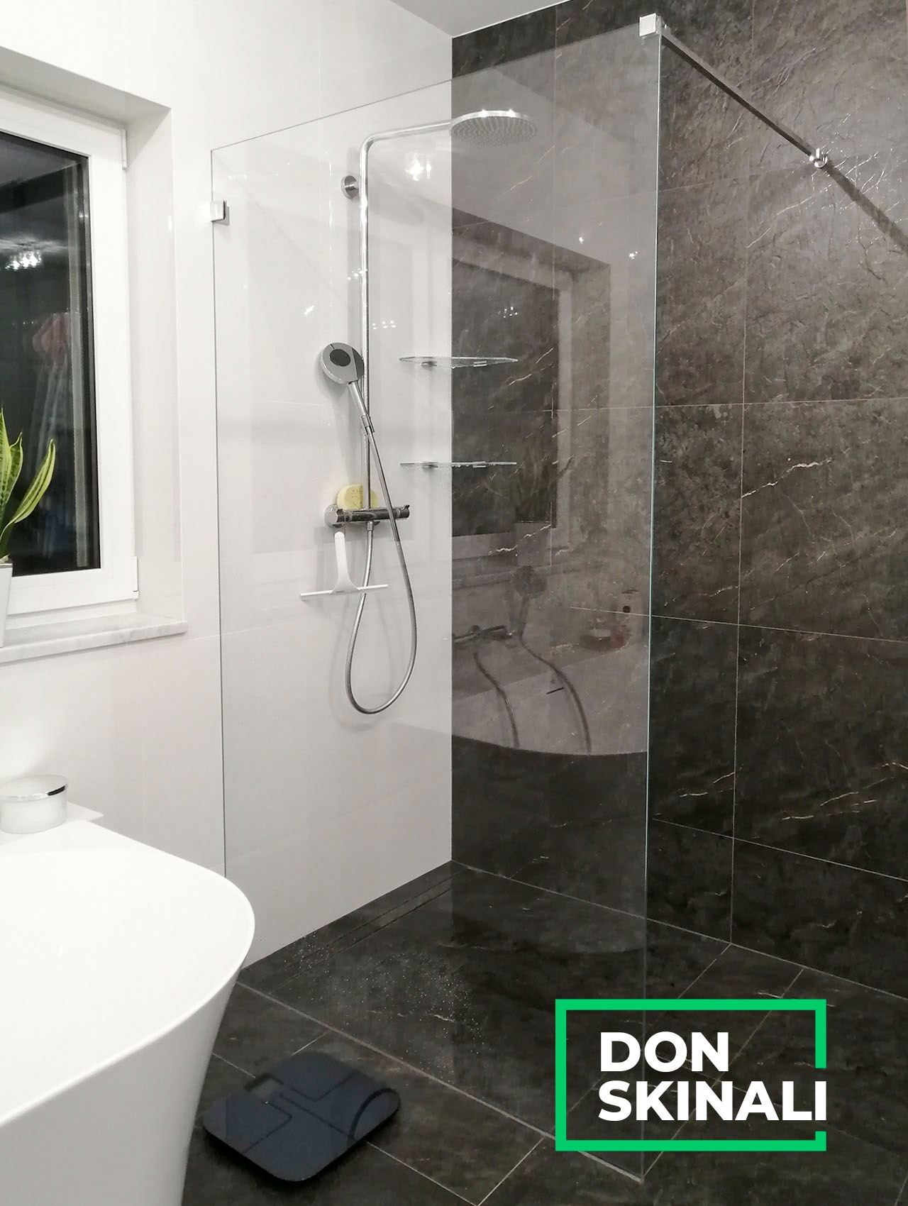Nowoczesna kabina prysznicowa z pojedynczą taflą szkła, chromowaną armaturą i półkami, na tle ciemnych płytek imitujących marmur.