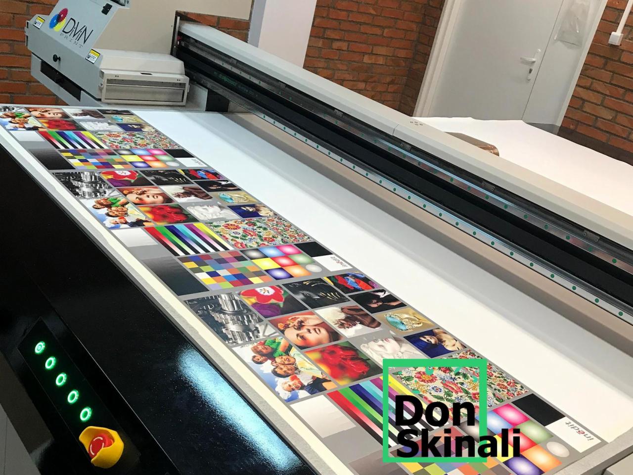 Wielkoformatowa drukarka drukująca próbnik kolorów i wzorów na materiale, widoczny panel sterowania z zielonymi diodami i czerwonym przyciskiem awaryjnym.