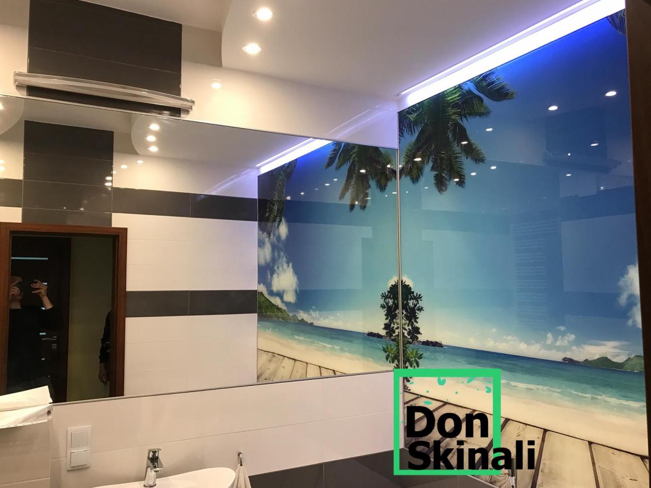 Szklana ściana z motywem plaży z palmami w łazience, odbicie w lustrze, logo firmy w prawym dolnym rogu.