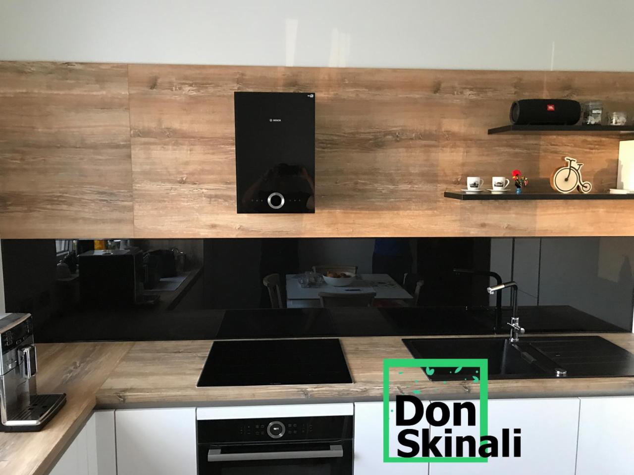 Nowoczesna kuchnia z czarnym panelem szklanym nad blatem, drewnopodobne szafki wiszące, białe szafki dolne, ekspres do kawy po lewej, płyta indukcyjna i piekarnik w zabudowie.