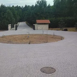 Okrągły podjazd z szarej kostki brukowej z centralnym klombem ziemi, w tle brama wjazdowa i las, widoczna przenośna toaleta.