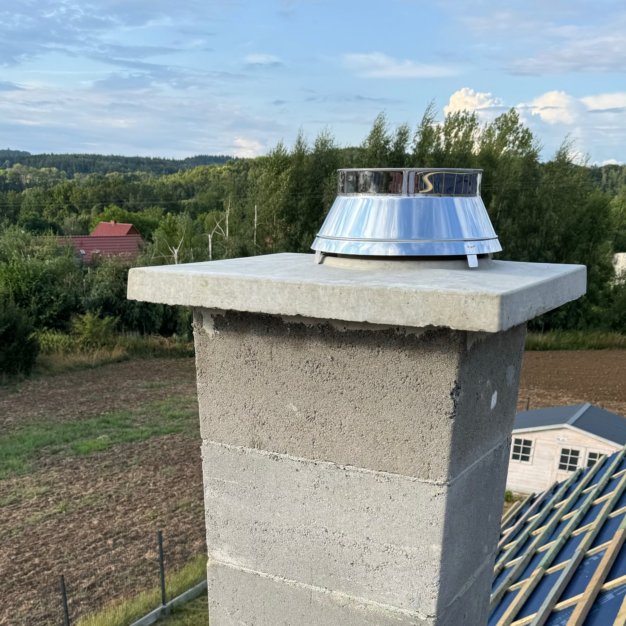 Betonowy komin z błyszczącym, metalowym daszkiem na tle pól i lasów, widok z dachu w trakcie budowy.