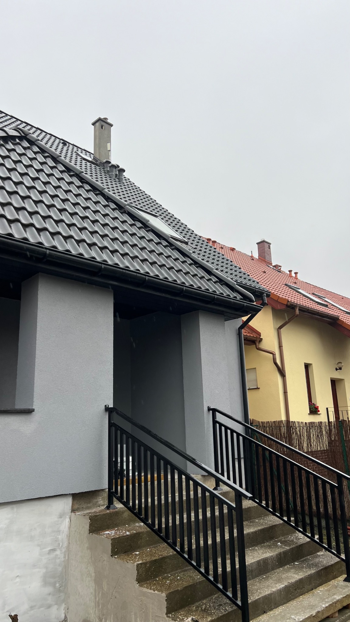 Szary dom z czarnym dachem i schodami z czarną balustradą, widok z dołu w pochmurny dzień.