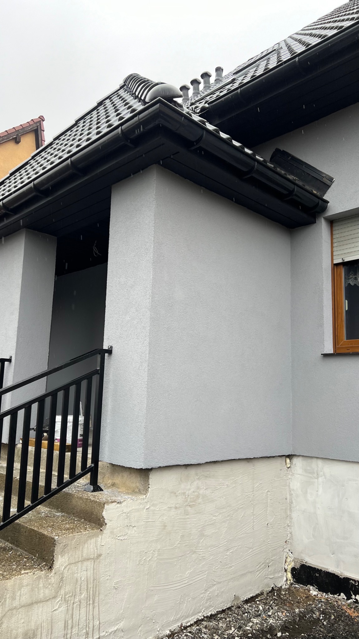 Odnowiona elewacja domu z szarą strukturą tynku, czarnym dachem z kominami i częściowo pomalowanym fundamentem przy schodach wejściowych.