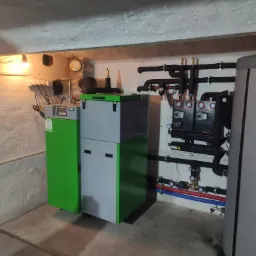 Nowoczesny, dwuczęściowy piec na paliwo stałe w kolorach szaro-zielonych, z podłączonymi rurami instalacji grzewczej i zbiornikiem akumulacyjnym w kotłowni o surowych ścianach.