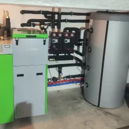 Szaro-zielony piec z podłączonym zbiornikiem akumulacyjnym, licznymi rurami w izolacji, manometrami i zaworami, zainstalowany w pomieszczeniu technicznym.