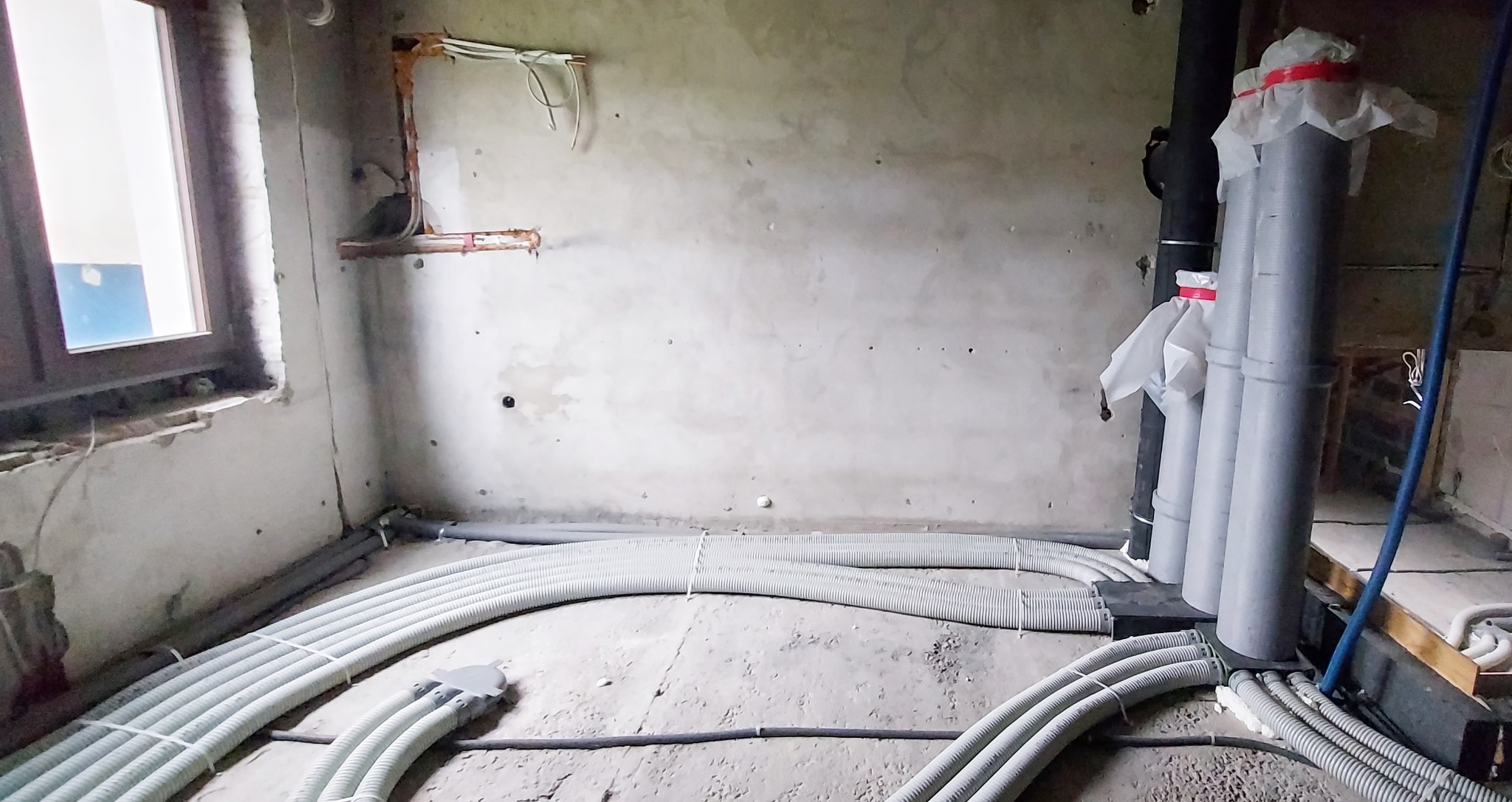 Wnętrze w trakcie remontu z widocznymi rurami instalacji grzewczej i pionami kanalizacyjnymi, ściany w stanie surowym, okno z widokiem na zewnątrz.