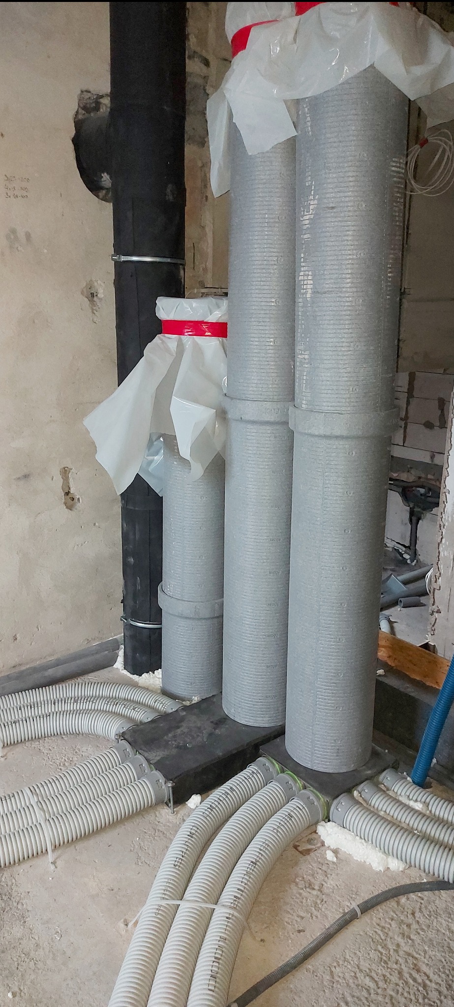 Szare rury kanalizacyjne pionowe i poziome, zabezpieczone folią, w trakcie montażu w surowym wnętrzu budynku.