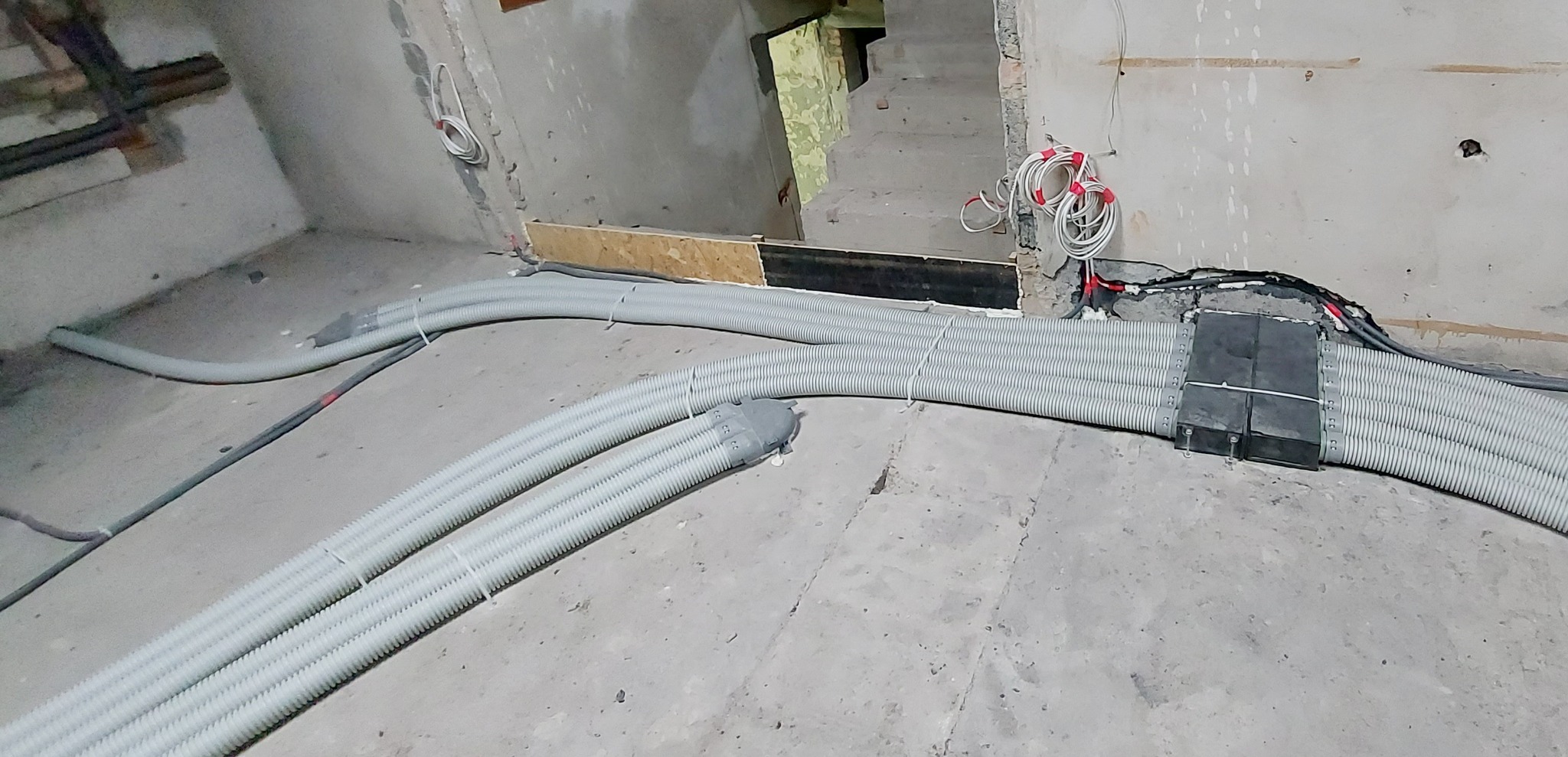Szare, karbowane rury instalacyjne ułożone na surowym betonowym podłożu w trakcie prac instalacyjnych, widoczne przewody elektryczne i fragment schodów w tle