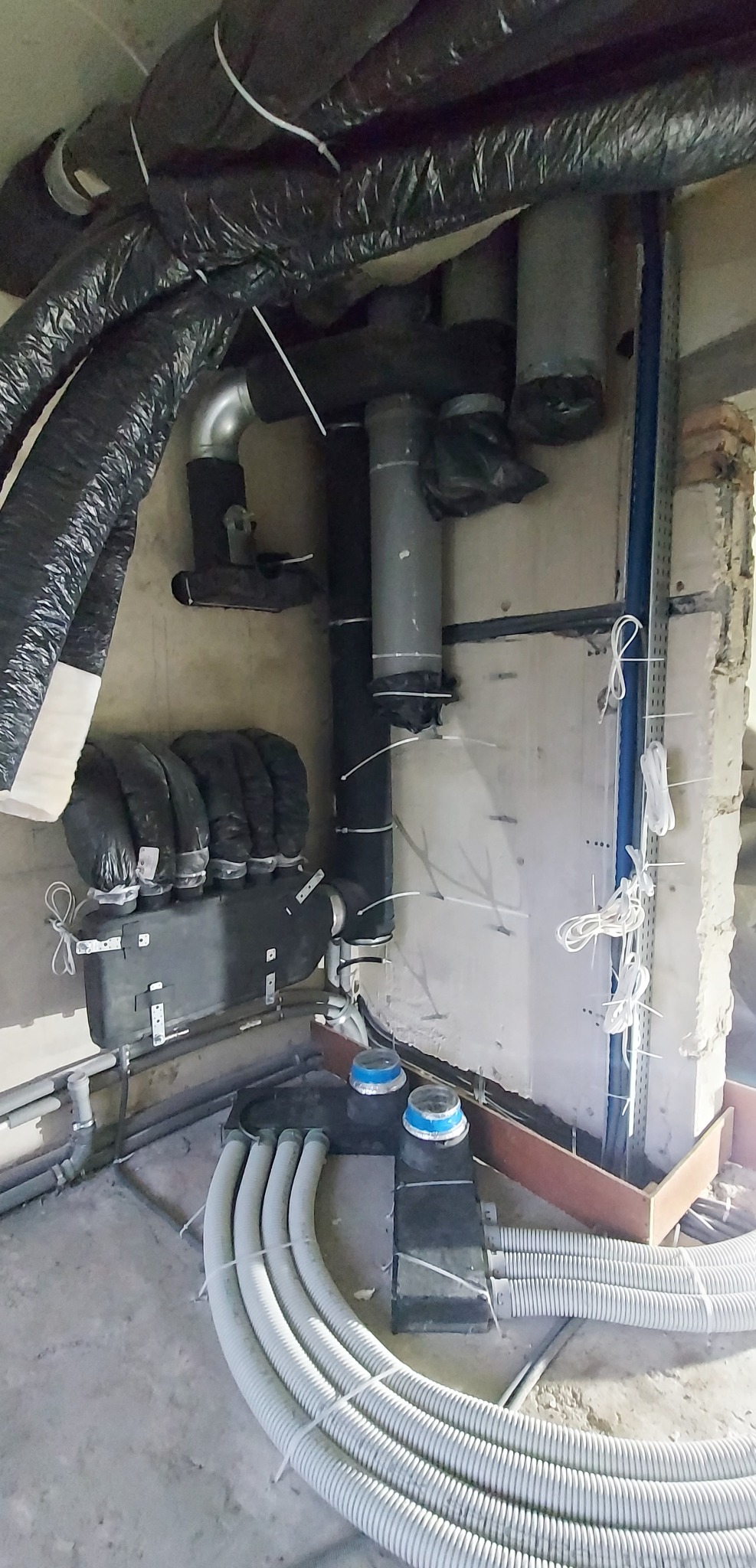 Wnętrze w trakcie montażu systemu rekuperacji: widoczne czarne izolowane rury wentylacyjne pod sufitem, szare rury PCV przymocowane do ściany za pomocą opasek zaciskowych oraz białe elastyczne...