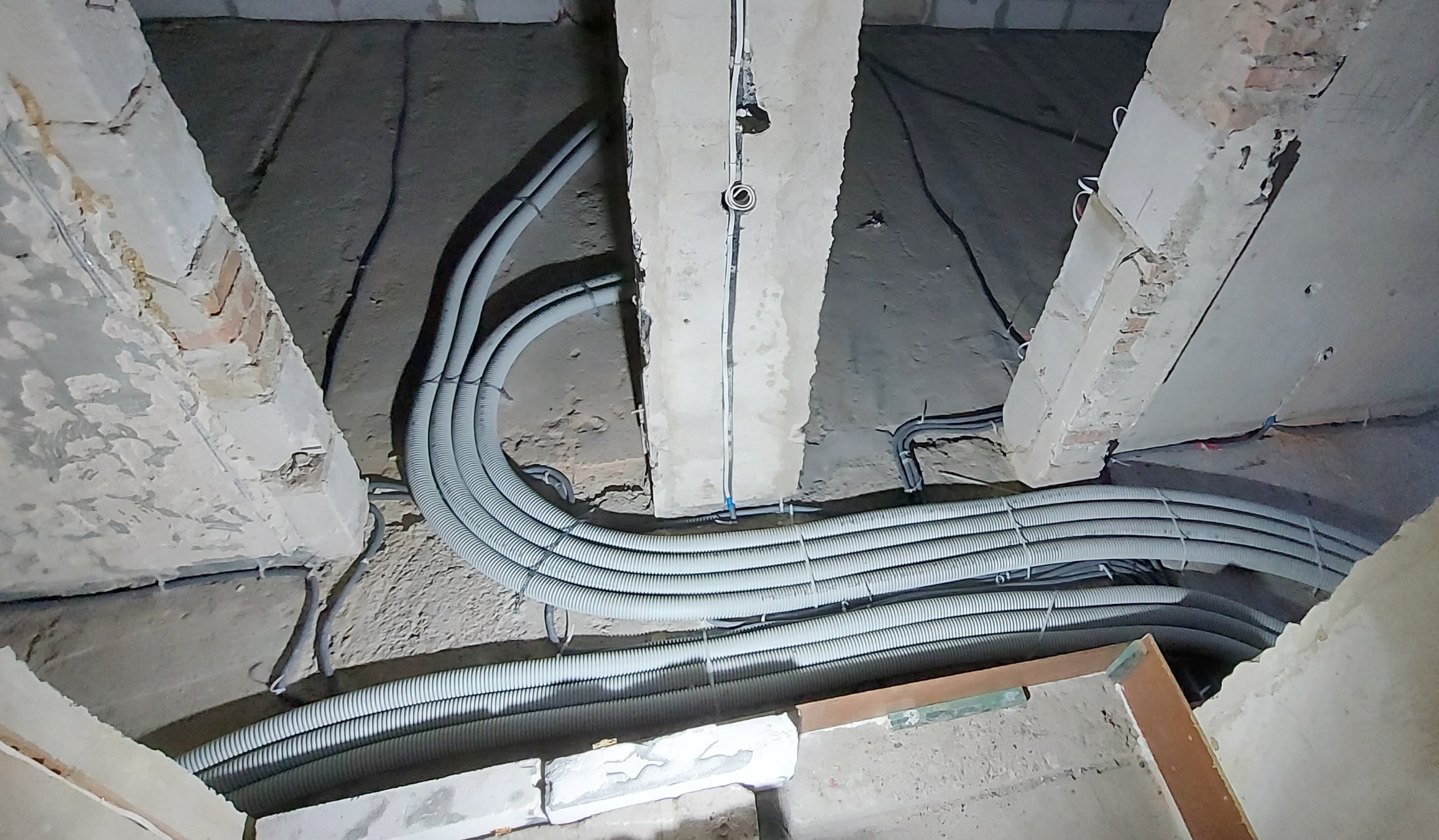 Szereg szarych, karbowanych rur instalacyjnych, prowadzonych pod betonowym stropem w surowym stanie, przymocowanych do niego za pomocą białych opasek zaciskowych.