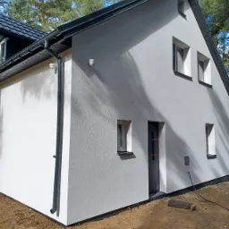 Nowoczesny dom z jasnoszarą elewacją o fakturze baranka, czarnym dachem i orynnowaniem, z widocznymi elementami wykończeniowymi przy ziemi, na tle lasu.