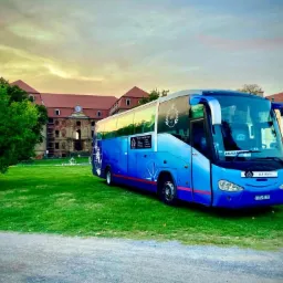 Niebieski autokar turystyczny zaparkowany na trawniku przed zabytkowym budynkiem z czerwoną dachówką i zegarem na fasadzie, widoczne logo firmy przewozowej na autokarze.