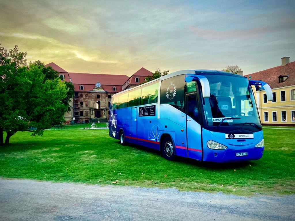 Niebieski autokar turystyczny zaparkowany na trawniku przed zabytkowym budynkiem z czerwoną dachówką i zegarem na fasadzie, widoczne logo firmy przewozowej na autokarze.
