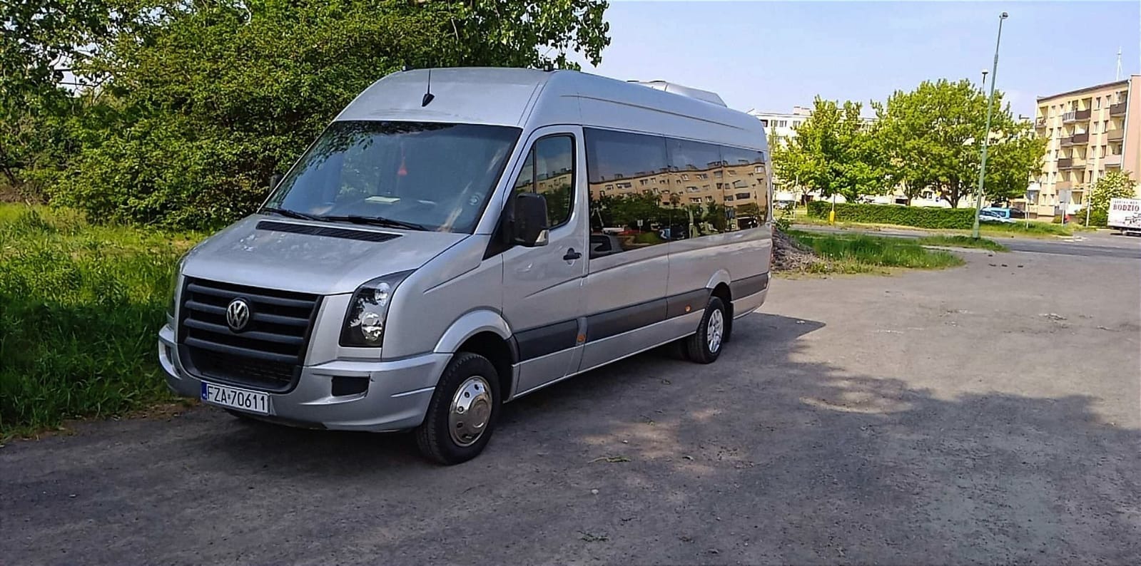 Srebrny bus Volkswagen Crafter z przyciemnionymi szybami, zaparkowany na placu w pobliżu zieleni i bloku mieszkalnego.