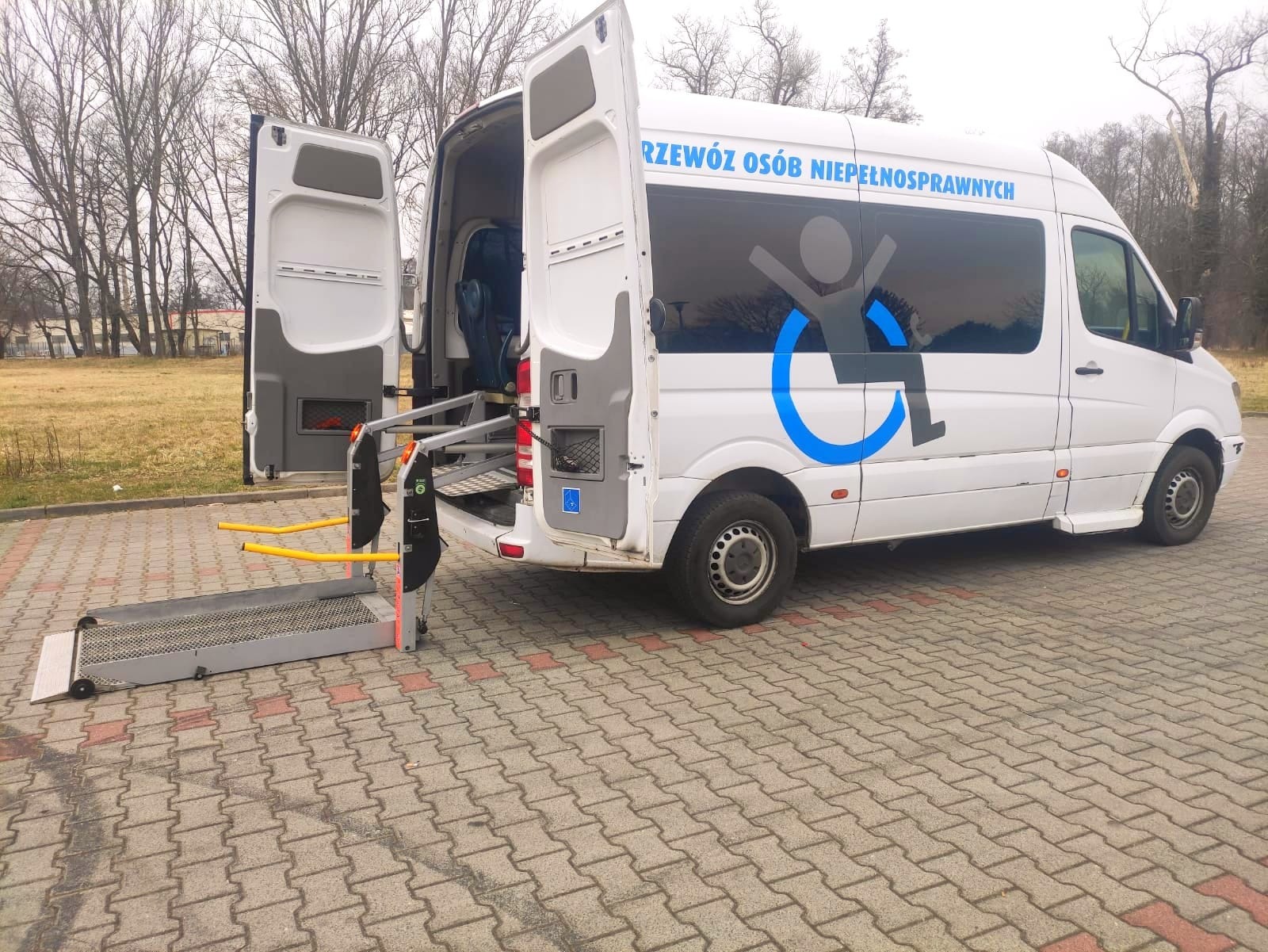 Biały bus z otwartymi drzwiami i wysuniętą rampą dla osób niepełnosprawnych, na boku niebieskie logo wózka inwalidzkiego i napis.