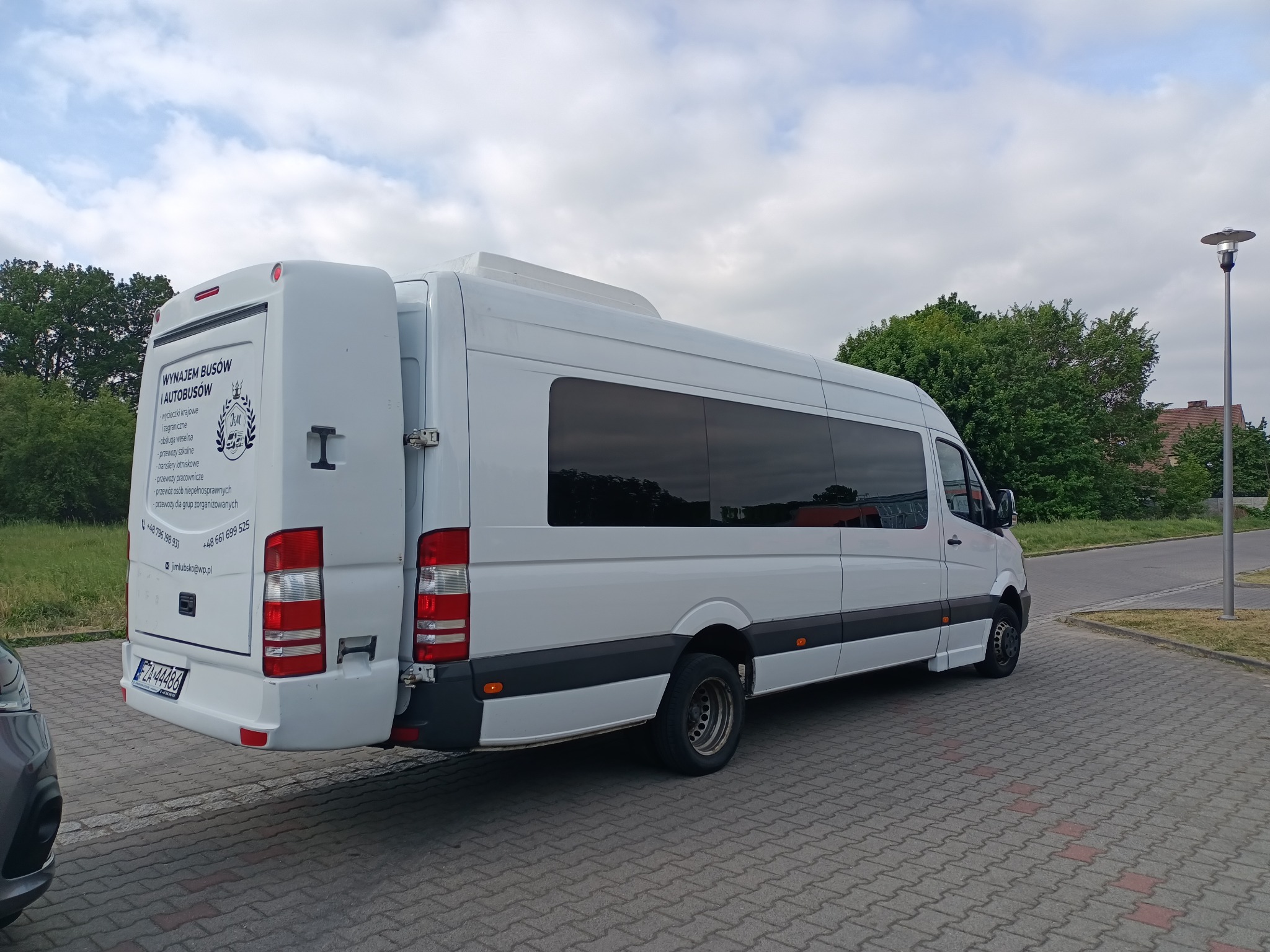 Biały bus z otwartymi drzwiami bagażnika, zaparkowany na kostce brukowej, z widocznym logo firmy oferującej wynajem busów i autobusów oraz numerem telefonu i adresem email na tylnych drzwiach.