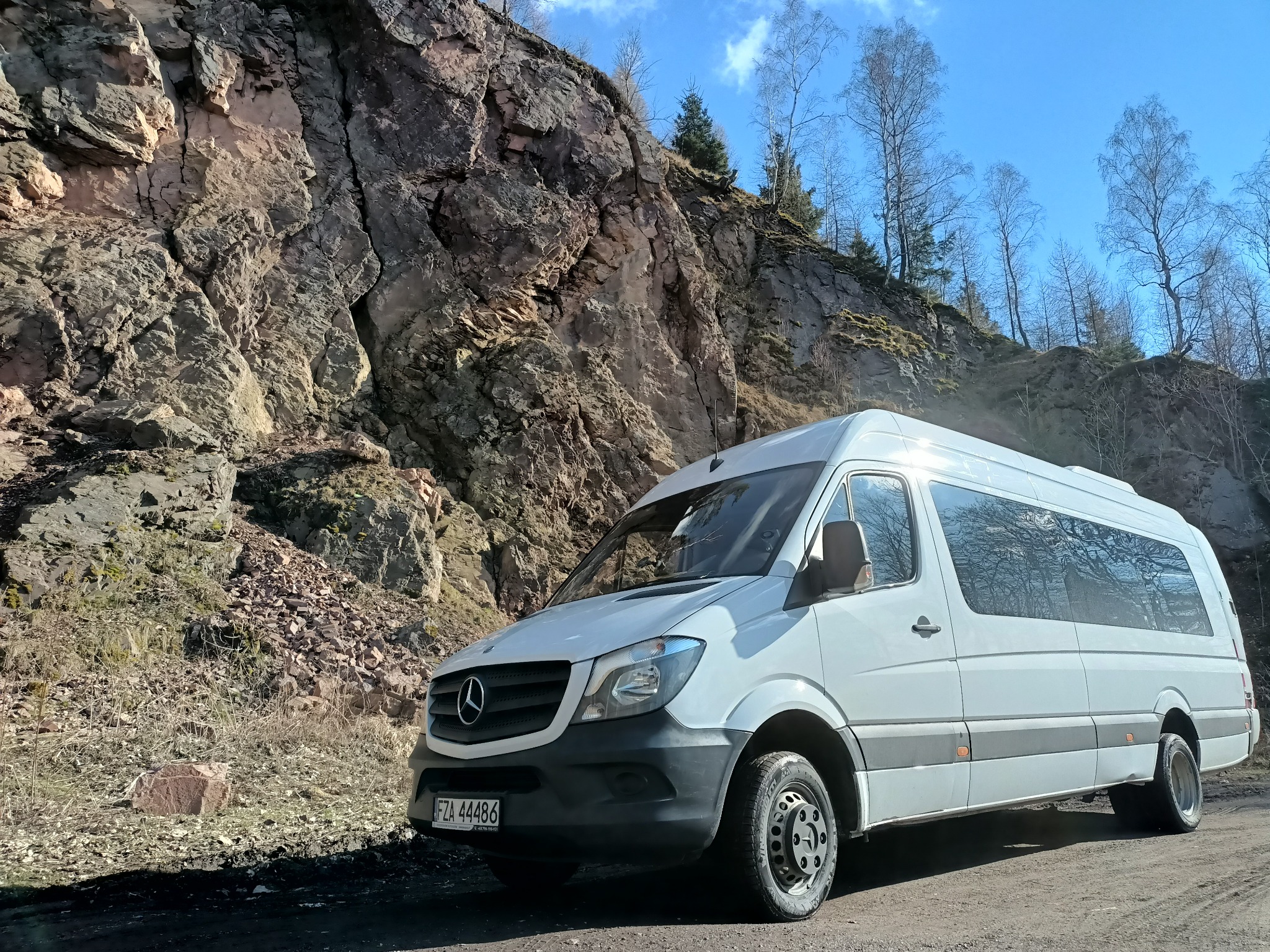 Biały bus Mercedes-Benz Sprinter z przyciemnianymi szybami pasażerskimi, zaparkowany na żwirowej drodze pod skalistym zboczem porośniętym drzewami bez liści, widoczne tablice rejestracyjne.