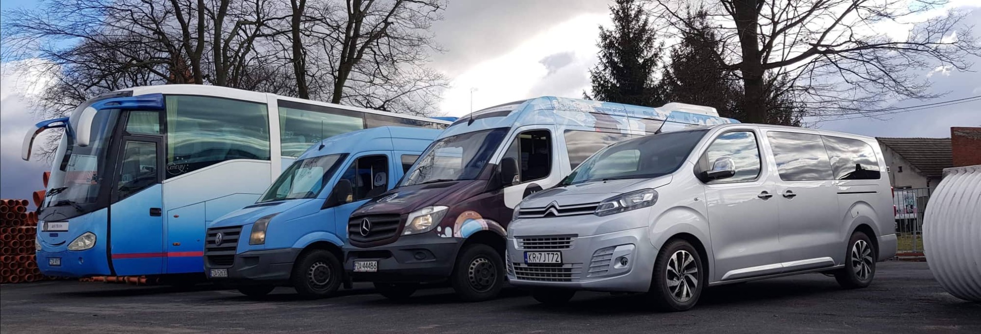 Flota pojazdów: autokar, busy i van, zaparkowane w rzędzie na tle drzew i pochmurnego nieba.