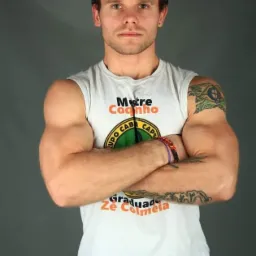 Mężczyzna w białym stroju Capoeira z logo, skrzyżowane ramiona, tatuaże na ramionach, sznurkowy pas, na szarym tle.