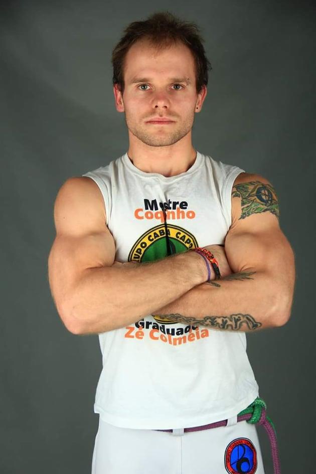 Mężczyzna w białym stroju Capoeira z logo, skrzyżowane ramiona, tatuaże na ramionach, sznurkowy pas, na szarym tle.