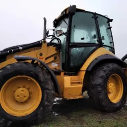 Żółta koparko-ładowarka CAT 434E z widocznymi śladami błota na oponach i karoserii, stojąca na grząskim terenie w pochmurny dzień.